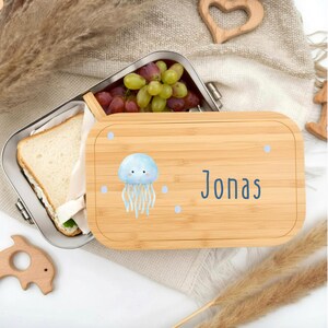 Pu&ograve; includere: Un portapranzo in bamb&ugrave; con interno in acciaio inossidabile, aperto per rivelare un panino e uva. Il coperchio presenta un'illustrazione di medusa blu, punti blu e il nome "Jonas" in lettere blu.