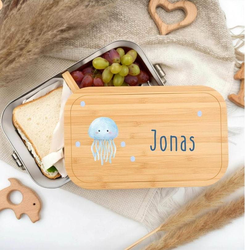 Pu&ograve; includere: Un portapranzo in bamb&ugrave; con interno in acciaio inossidabile, aperto per rivelare un panino e uva. Il coperchio presenta un'illustrazione di medusa blu, punti blu e il nome "Jonas" in lettere blu.