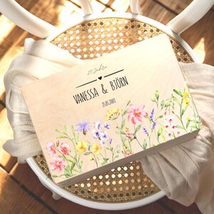 K&ouml;nnte beinhalten: Eine Holz-Andenkenbox mit floralen Aquarellkunstwerken. Die Box ist hellfarben mit den Namen "VANESSA & BJ&Ouml;RN" und dem Datum "25.05.2001" auf der Vorderseite. Die Box ist mit bunten Blumen verziert.