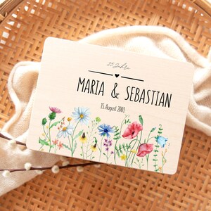 May include: A wooden plaque with the names "Maria & Sebastian" and the date "15. August 2001" in black lettering. The plaque is decorated with a watercolor floral border featuring red poppies, bluebells, and other wildflowers.