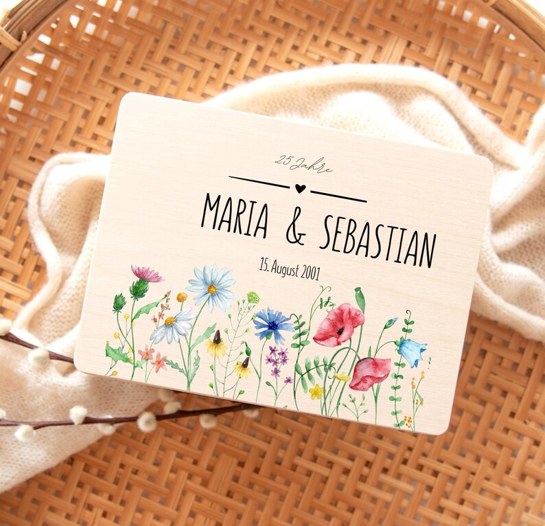May include: A wooden plaque with the names "Maria & Sebastian" and the date "15. August 2001" in black lettering. The plaque is decorated with a watercolor floral border featuring red poppies, bluebells, and other wildflowers.