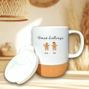 May include: A white ceramic mug with a cork base and a matching white lid. The mug features the text Omas Lieblinge above two gingerbread person illustrations, one with a candy cane and one with a red bow, labeled Larissa and Marie.