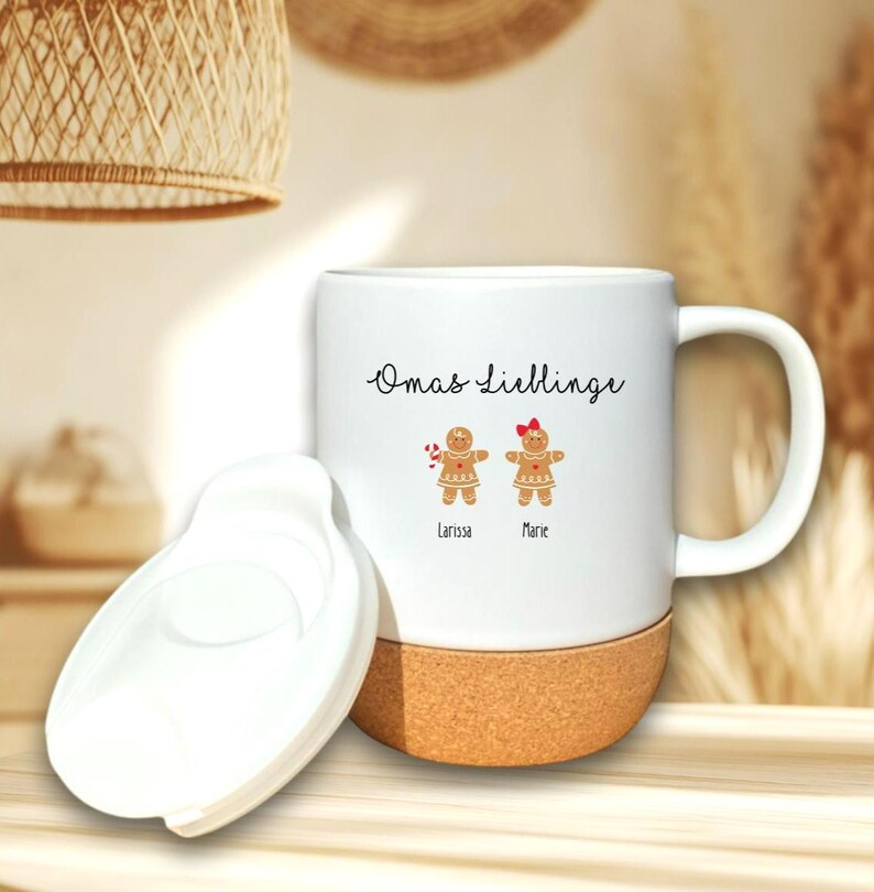 May include: A white ceramic mug with a cork base and a matching white lid. The mug features the text Omas Lieblinge above two gingerbread person illustrations, one with a candy cane and one with a red bow, labeled Larissa and Marie.