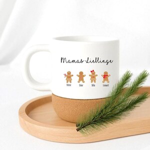 May include: A white ceramic mug with a cork base featuring the text Mamas Lieblinge and four gingerbread cookie illustrations labeled with the names Helene, Oskar, Bella, and Leonard, with one cookie holding a candy cane and one wearing a red bow.