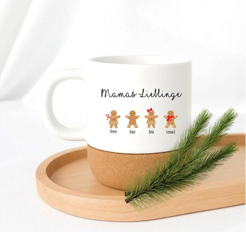 May include: A white ceramic mug with a cork base featuring the text Mamas Lieblinge and four gingerbread cookie illustrations labeled with the names Helene, Oskar, Bella, and Leonard, with one cookie holding a candy cane and one wearing a red bow.