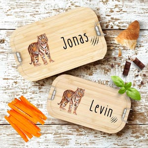 Pu&ograve; includere: Due portapranzo rettangolari in legno con chiusure in metallo. Ogni scatola presenta un'illustrazione di tigre e un nome: "Jonas" e "Levin". Le scatole sono su una superficie di legno imbiancato, con bastoncini di carote, un croissant e pezzi di cioccolato.