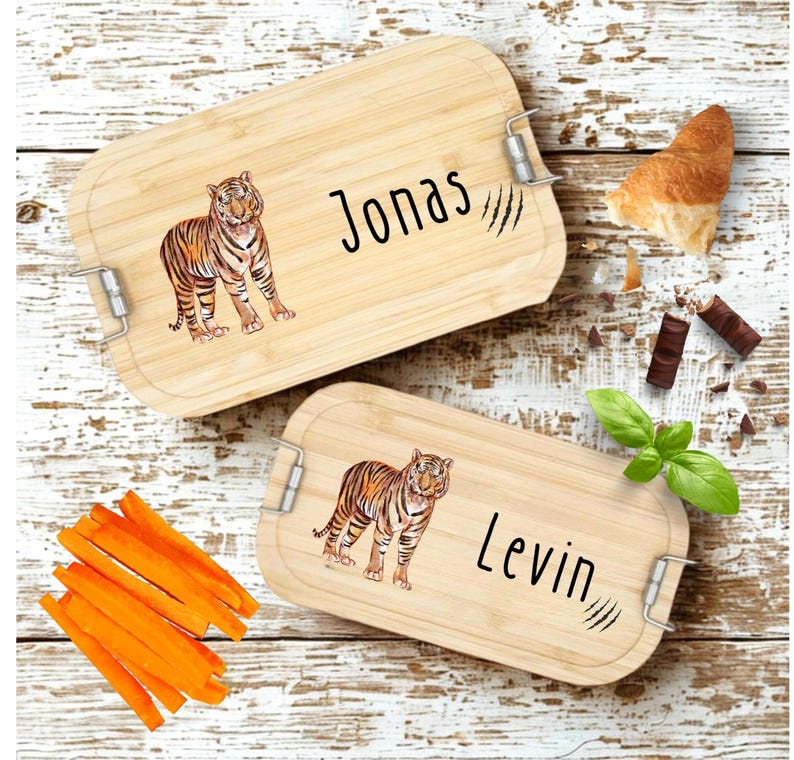 Pu&ograve; includere: Due portapranzo rettangolari in legno con chiusure in metallo. Ogni scatola presenta un'illustrazione di tigre e un nome: "Jonas" e "Levin". Le scatole sono su una superficie di legno imbiancato, con bastoncini di carote, un croissant e pezzi di cioccolato.