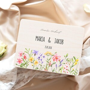 May include: A wooden wedding keepsake box with the names "MARIA & JAKOB" and the date "25.05.2026" printed on it. The box is decorated with a watercolor floral design featuring pink, yellow, and purple flowers.