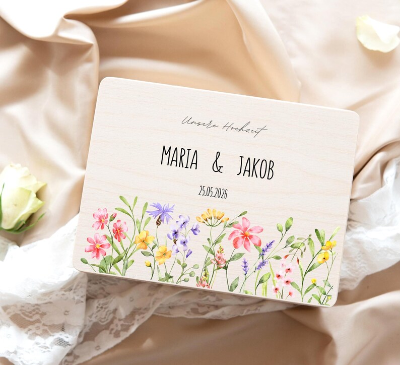 May include: A wooden wedding keepsake box with the names "MARIA & JAKOB" and the date "25.05.2026" printed on it. The box is decorated with a watercolor floral design featuring pink, yellow, and purple flowers.