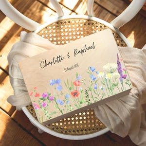 May include: A wooden box with a floral design and the names "Charlotte & Raphael" and the date "15. August 2026" in black script. The box is decorated with watercolor flowers in shades of red, purple, blue, and white.