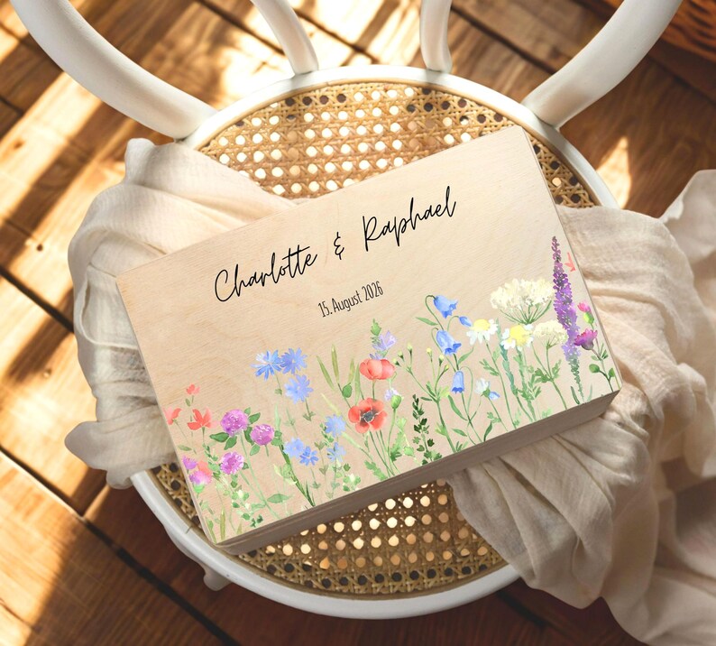 May include: A wooden box with a floral design and the names "Charlotte & Raphael" and the date "15. August 2026" in black script. The box is decorated with watercolor flowers in shades of red, purple, blue, and white.