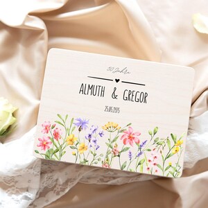 K&ouml;nnte beinhalten: Eine rechteckige, helle Holzbox mit einem aufgedruckten Blumenmuster am unteren Rand, bestehend aus rosa, gelben und violetten Bl&uuml;ten. Der Text lautet 50 Jahre, ALMUTH & GREGOR und 25.05.1975, getrennt durch ein kleines schwarzes Herz.