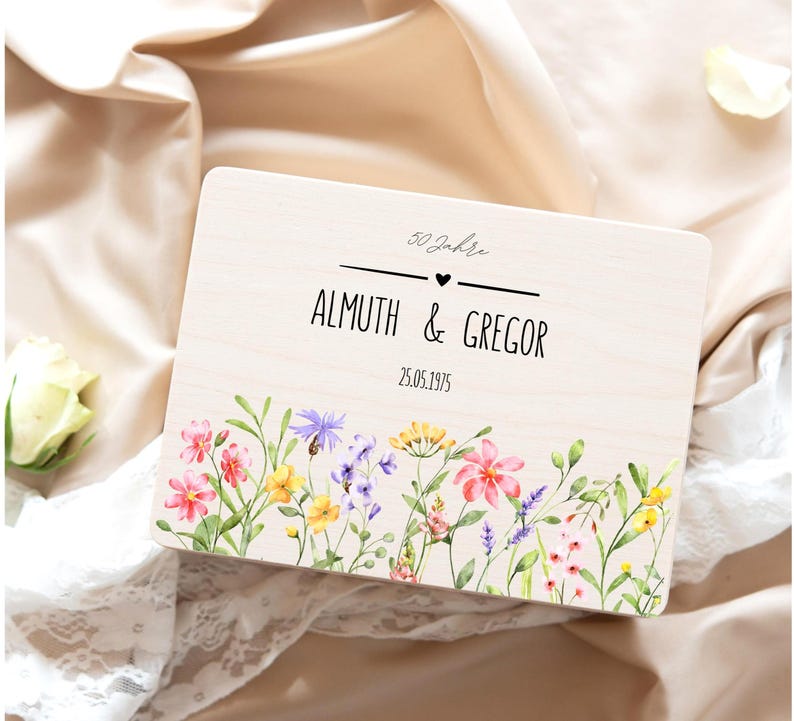 K&ouml;nnte beinhalten: Eine rechteckige, helle Holzbox mit einem aufgedruckten Blumenmuster am unteren Rand, bestehend aus rosa, gelben und violetten Bl&uuml;ten. Der Text lautet 50 Jahre, ALMUTH & GREGOR und 25.05.1975, getrennt durch ein kleines schwarzes Herz.