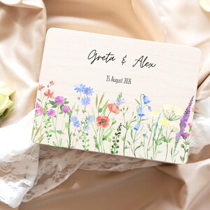 May include: A wooden box with a floral watercolor design. The box is rectangular with the names "Greta & Alex" and the date "15. August 2026" printed on the top. The floral design includes red poppies, bluebells, and purple lavender.