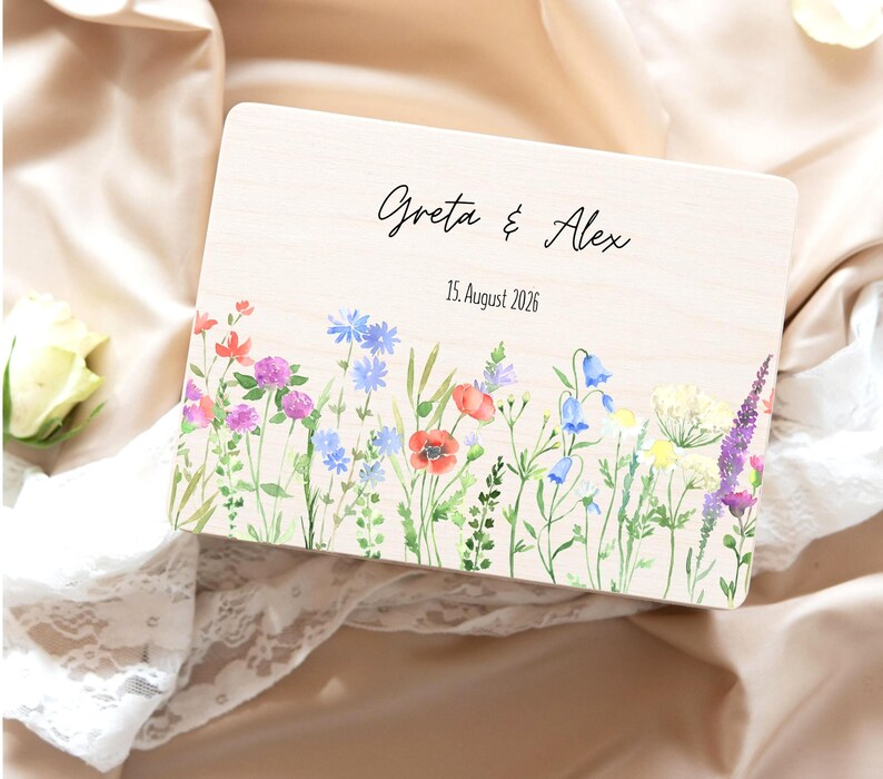 May include: A wooden box with a floral watercolor design. The box is rectangular with the names "Greta & Alex" and the date "15. August 2026" printed on the top. The floral design includes red poppies, bluebells, and purple lavender.
