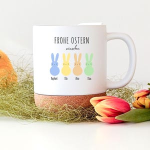 Könnte beinhalten: Weißer Keramikbecher mit Korkboden, mit dem Text "Frohe Ostern wünschen" und vier pastellfarbenen Hasenillustrationen. Jeder Hase hat einen Namen darunter. Der Becher steht auf einem Bett aus grünem Stroh, mit einem gelben Küken und Tulpen.