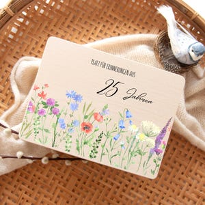 Op de afbeelding: Een houten plaat met de tekst "PLATZ F&Uuml;R ERINNERUNGEN AUS 25 Jahren" en een aquarel bloemenrand. De bloemen omvatten rode klaprozen, blauwe korenbloemen en paarse lavendel. Een decoratieve vogel zit in een nest.