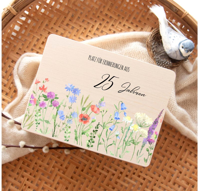 Op de afbeelding: Een houten plaat met de tekst "PLATZ F&Uuml;R ERINNERUNGEN AUS 25 Jahren" en een aquarel bloemenrand. De bloemen omvatten rode klaprozen, blauwe korenbloemen en paarse lavendel. Een decoratieve vogel zit in een nest.
