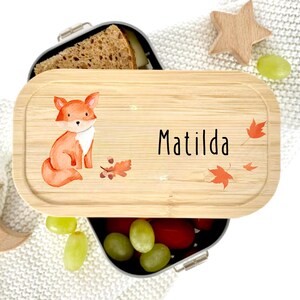 Pu&ograve; includere: Un portapranzo in bamb&ugrave; con un'illustrazione di volpe ad acquerello e il nome "Matilda" in lettere nere. Il coperchio presenta foglie autunnali e una piccola stella di legno. Il portapranzo &egrave; aperto, rivelando cibo.