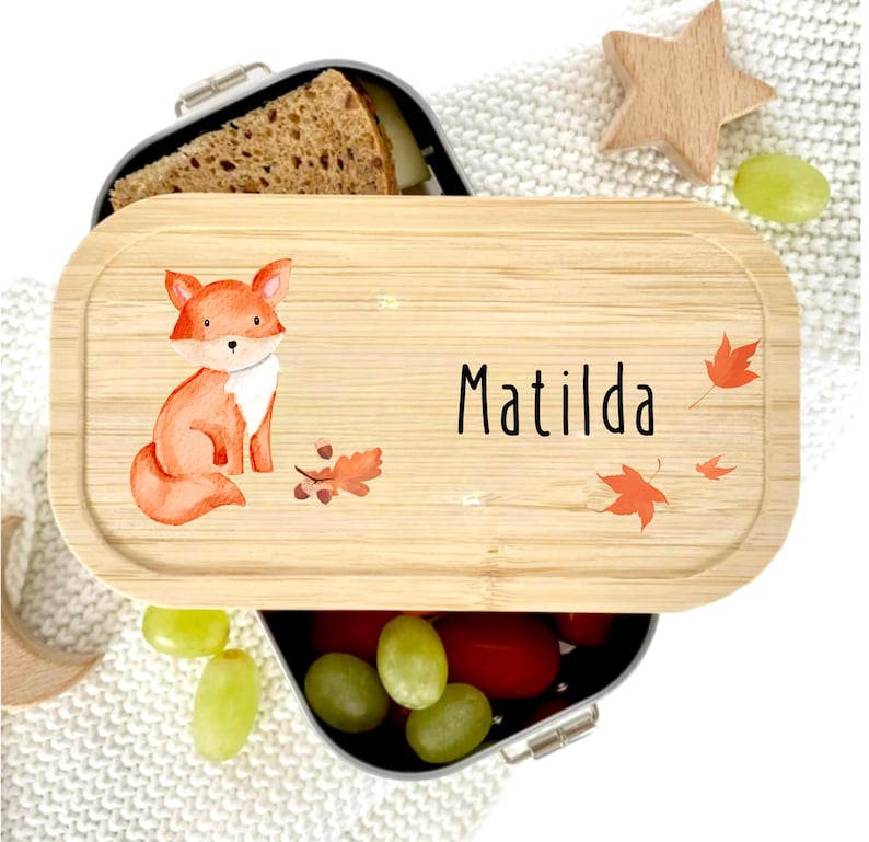 Pu&ograve; includere: Un portapranzo in bamb&ugrave; con un'illustrazione di volpe ad acquerello e il nome "Matilda" in lettere nere. Il coperchio presenta foglie autunnali e una piccola stella di legno. Il portapranzo &egrave; aperto, rivelando cibo.
