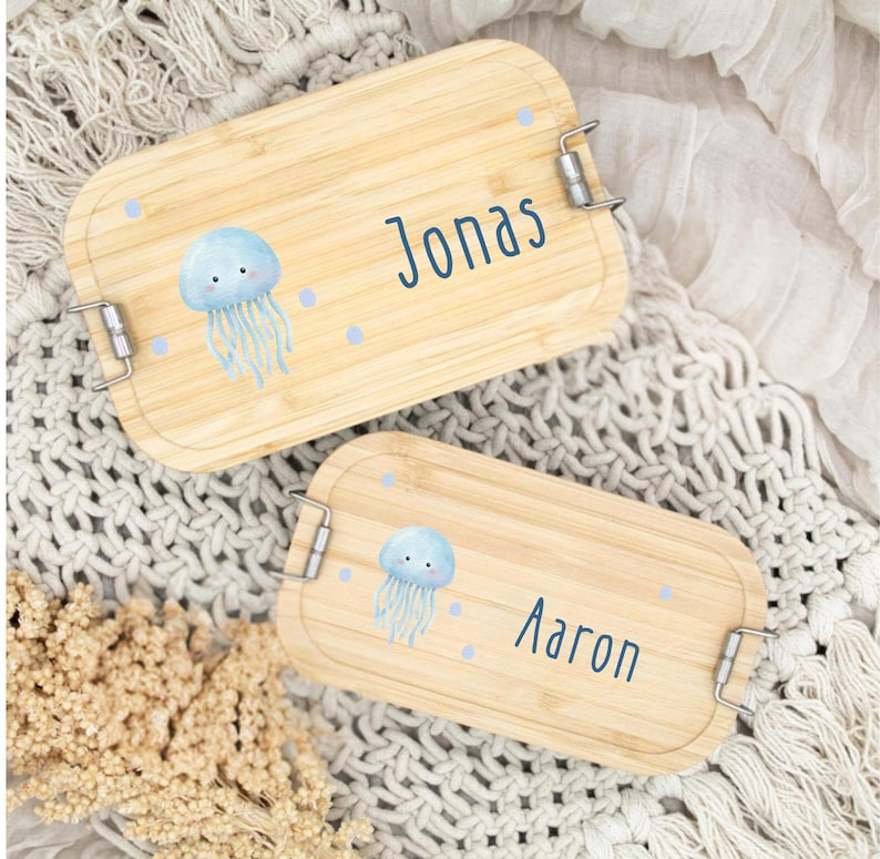 Pu&ograve; includere: Due portapranzo rettangolari in bamb&ugrave;. Ogni scatola ha una chiusura argentata ed &egrave; decorata con un'illustrazione di medusa blu e il nome "Jonas" o "Aaron" in blu. Sono presenti anche pois azzurri.