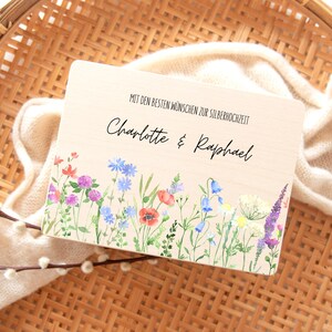 Puede incluir: Una placa de madera con el texto "MIT DEN BESTEN W&Uuml;NSCHEN ZUR SILBERHOCHZEIT" y los nombres "Charlotte & Raphael". El borde inferior presenta un dise&ntilde;o floral en acuarela con flores rojas, azules y moradas.