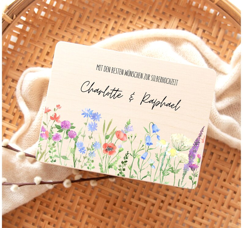 Puede incluir: Una placa de madera con el texto "MIT DEN BESTEN W&Uuml;NSCHEN ZUR SILBERHOCHZEIT" y los nombres "Charlotte & Raphael". El borde inferior presenta un dise&ntilde;o floral en acuarela con flores rojas, azules y moradas.