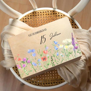 Op de afbeelding: Een houten kist met een bloemenontwerp en de tekst "Platz f&uuml;r Erinnerungen aus 15 Jahren." De kist heeft een aquarel illustratie van wilde bloemen in tinten rood, blauw, paars en wit. De kist rust op een gevlochten stoel.