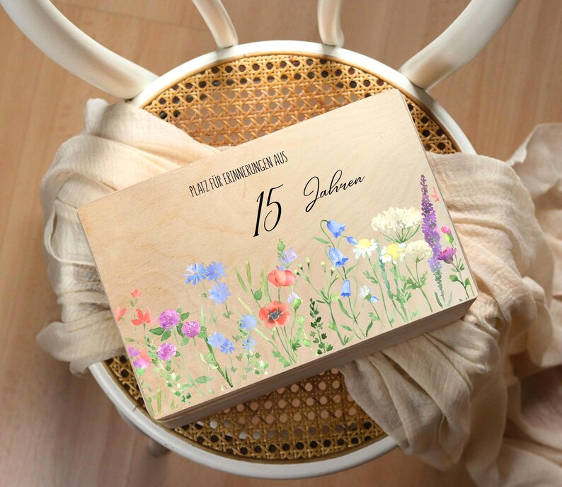 Op de afbeelding: Een houten kist met een bloemenontwerp en de tekst "Platz f&uuml;r Erinnerungen aus 15 Jahren." De kist heeft een aquarel illustratie van wilde bloemen in tinten rood, blauw, paars en wit. De kist rust op een gevlochten stoel.