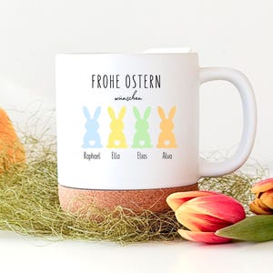 Könnte beinhalten: Weißer Keramikbecher mit Henkel, mit dem Text "FROHE OSTERN wünschen" und vier bunten Hasenillustrationen. Unter den Hasen stehen die Namen Raphael, Ella, Elias und Alva. Der Becher steht auf einem Korksockel.