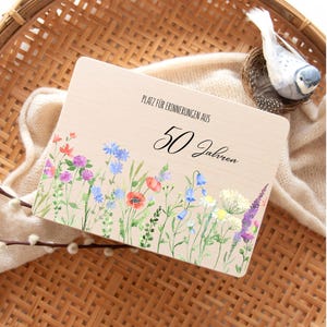 Op de afbeelding: Houten plaat met de tekst "PLATZ F&Uuml;R ERINNERUNGEN AUS 50 Jahren" en een aquarel bloemenrand. De bloemen zijn onder meer rode klaprozen, blauwe klokjes en paarse lavendel. Een decoratieve vogel zit in een nest.