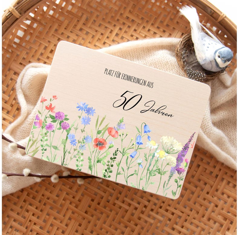 Op de afbeelding: Houten plaat met de tekst "PLATZ F&Uuml;R ERINNERUNGEN AUS 50 Jahren" en een aquarel bloemenrand. De bloemen zijn onder meer rode klaprozen, blauwe klokjes en paarse lavendel. Een decoratieve vogel zit in een nest.
