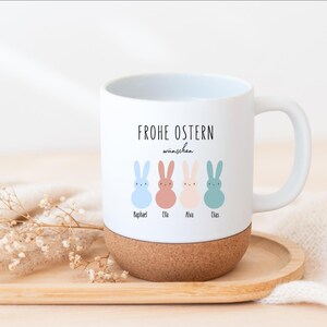 Könnte beinhalten: Weiße Keramik-Tasse mit Korkboden, mit dem Text "FROHE OSTERN wünschen" und vier pastellfarbenen Hasen-Illustrationen. Jeder Hase hat einen Namen darunter. Die Tasse steht auf einem Holztablett.
