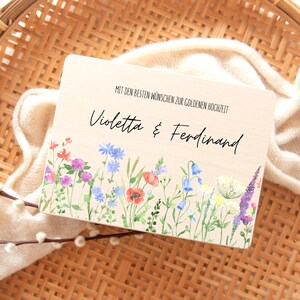Puede incluir: Placa de madera con el texto "Violetta & Ferdinand" y "MIT DEN BESTEN W&Uuml;NSCHEN ZUR GOLDENEN HOCHZEIT". La placa presenta un borde floral de acuarela con amapolas rojas, campanillas azules y lavanda p&uacute;rpura. El fondo es de color beige claro.