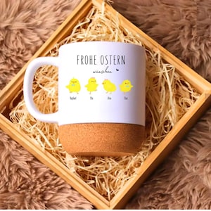 Könnte beinhalten: Weißer Keramikbecher mit Korkboden, mit dem Text "FROHE OSTERN wünschen" und Illustrationen von vier gelben Küken. Der Becher wird in einer Holzkiste mit Stroh präsentiert, vor einem braunen, strukturierten Hintergrund.