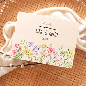 K&ouml;nnte beinhalten: Holzschild mit den Namen "LENA & PHILIPP" und dem Datum "25.05.2016" in Schwarz. Es hat einen floralen Rand mit rosa, gelben und lila Blumen. Der Text "10 Jahre" steht &uuml;ber den Namen.