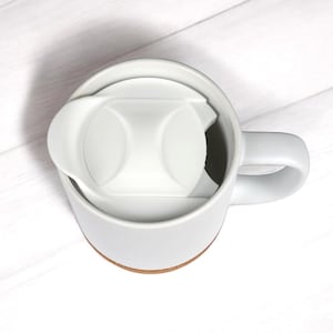 May include: A white ceramic mug featuring a matching white plastic lid with a contoured design. The mug has a smooth, cylindrical body and a sturdy side handle, with a visible terracotta-colored base rim, viewed from a high-angle perspective.