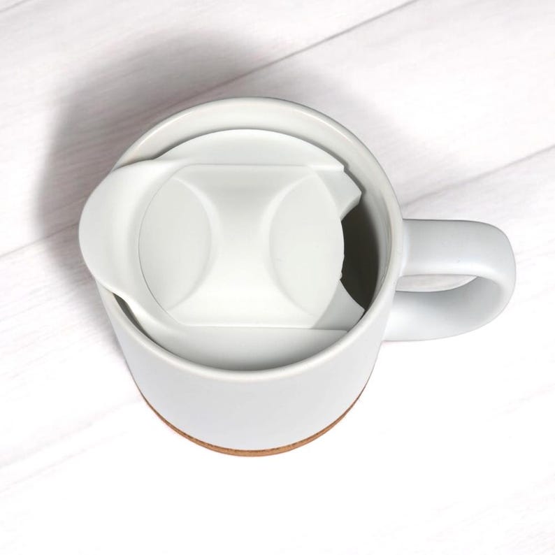 May include: A white ceramic mug featuring a matching white plastic lid with a contoured design. The mug has a smooth, cylindrical body and a sturdy side handle, with a visible terracotta-colored base rim, viewed from a high-angle perspective.