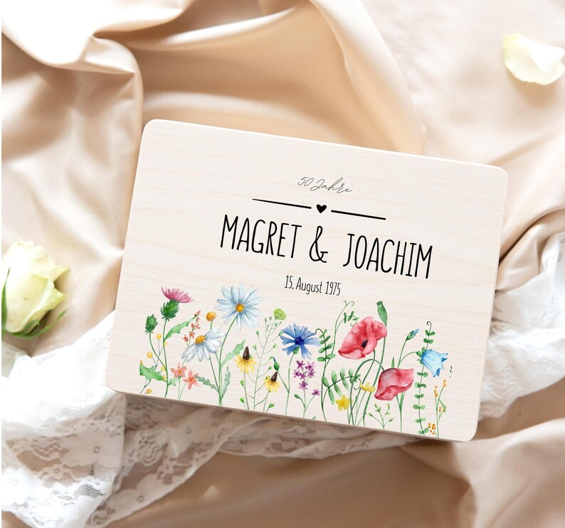 May include: A light-colored wooden box with the names "Magret & Joachim" and the date "15. August 1975" printed on it. The box is decorated with a watercolor floral design. The text "50 Jahre" is also printed on the box.