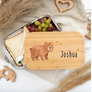 Pu&ograve; includere: Un portapranzo in bamb&ugrave; con un'illustrazione di un orso marrone e il nome "Joshua". Il portapranzo &egrave; aperto, rivelando un contenitore in acciaio inossidabile con un panino e uva. Il coperchio ha tre illustrazioni di api gialle.