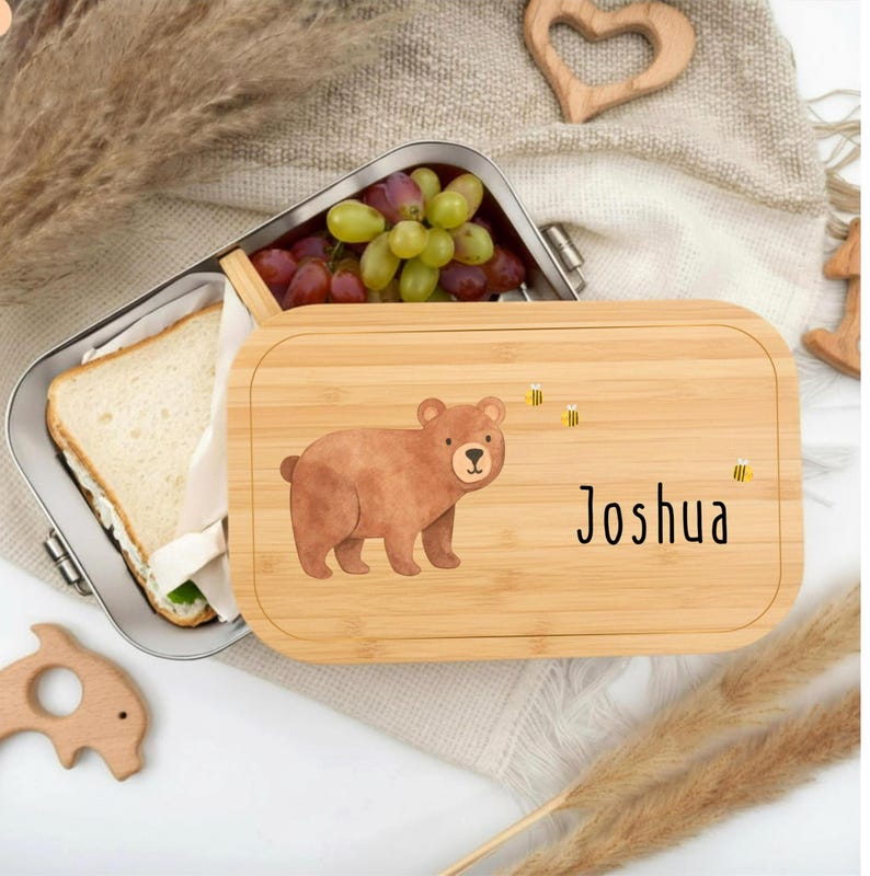 Pu&ograve; includere: Un portapranzo in bamb&ugrave; con un'illustrazione di un orso marrone e il nome "Joshua". Il portapranzo &egrave; aperto, rivelando un contenitore in acciaio inossidabile con un panino e uva. Il coperchio ha tre illustrazioni di api gialle.