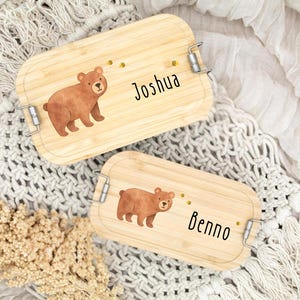 Pu&ograve; includere: Due portapranzo rettangolari in legno con chiusure in metallo. Ogni scatola presenta un'illustrazione di orso marrone e un nome in grafia nera: "Joshua" e "Benno". Sono presenti anche piccole grafiche di api gialle.