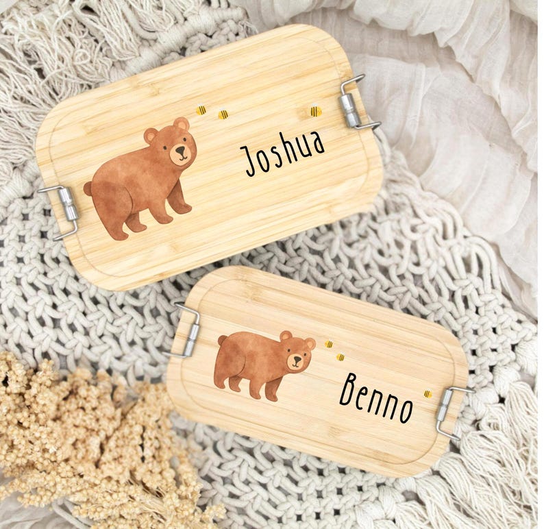 Pu&ograve; includere: Due portapranzo rettangolari in legno con chiusure in metallo. Ogni scatola presenta un'illustrazione di orso marrone e un nome in grafia nera: "Joshua" e "Benno". Sono presenti anche piccole grafiche di api gialle.