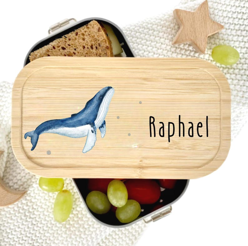 Pu&ograve; includere: Un portapranzo in bamb&ugrave; con un'illustrazione di balena blu e il nome "Raphael" in nero. Il portapranzo &egrave; aperto, rivelando cibo. Una stella di legno e dell'uva sono vicine.
