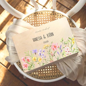 May include: A wooden guest book with floral watercolor art. The book features the names "VANESSA & BJ&Ouml;RN" and the date "25.05.2026". The book is decorated with colorful flowers in pink, yellow, purple, and white.