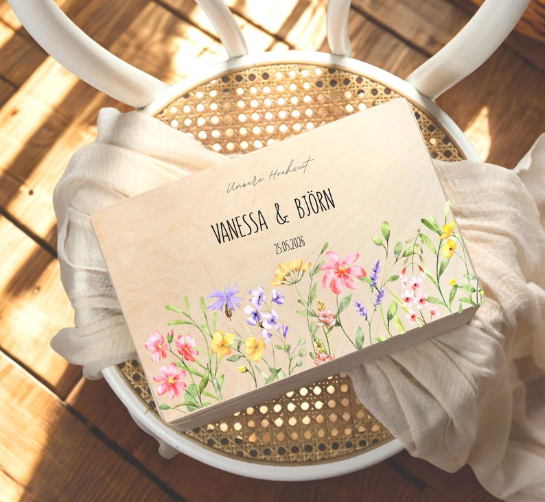 May include: A wooden guest book with floral watercolor art. The book features the names "VANESSA & BJ&Ouml;RN" and the date "25.05.2026". The book is decorated with colorful flowers in pink, yellow, purple, and white.