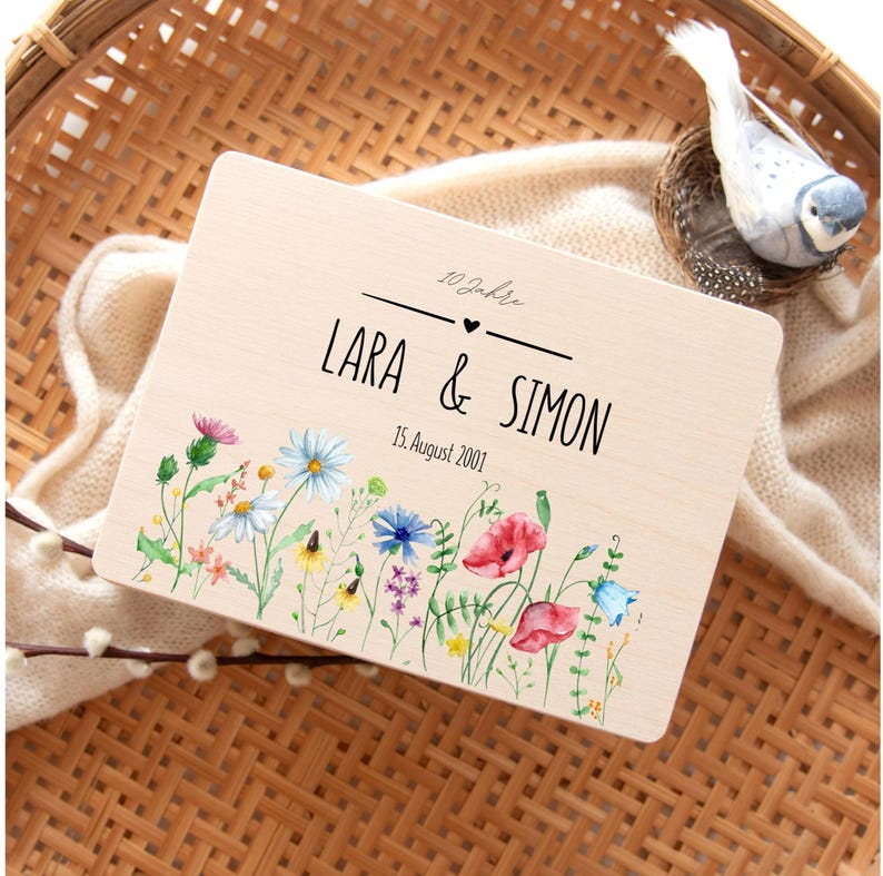 May include: A wooden plaque with the names "LARA & SIMON" and the date "15. August 2001" in black lettering. The plaque is decorated with a watercolor floral border featuring red poppies, blue cornflowers, and other wildflowers.