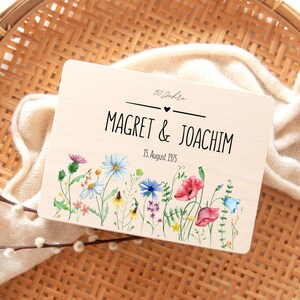 May include: A wooden plaque with the names "Magret & Joachim" and the date "15. August 1975" in black lettering. The plaque is decorated with a watercolor floral design featuring colorful wildflowers. The text "50 Jahre" is at the top.
