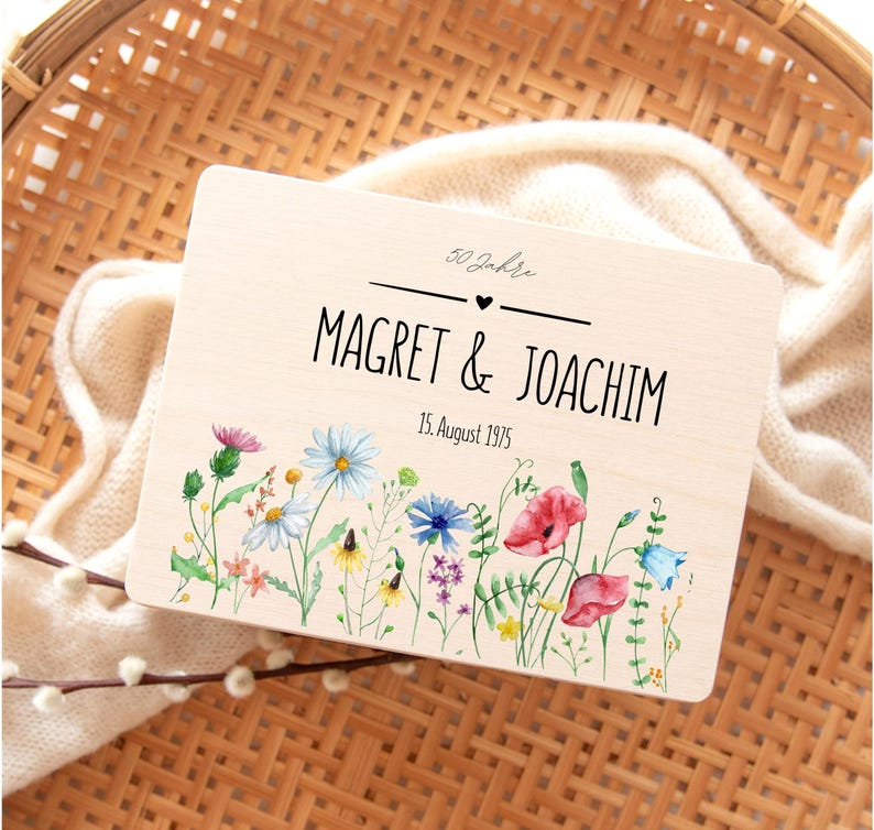 May include: A wooden plaque with the names "Magret & Joachim" and the date "15. August 1975" in black lettering. The plaque is decorated with a watercolor floral design featuring colorful wildflowers. The text "50 Jahre" is at the top.