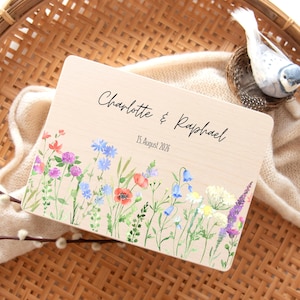 May include: A wooden sign with the names "Charlotte & Raphael" and the date "15. August 2026" in elegant script. The sign is decorated with a watercolor floral border featuring colorful wildflowers.